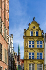 Yellow ornate facade with decorative details and tall windows beside classic brick buildings, St. Mary's Church spire rising behind under a bright blue sky in Gdańsk's old town © photoopus