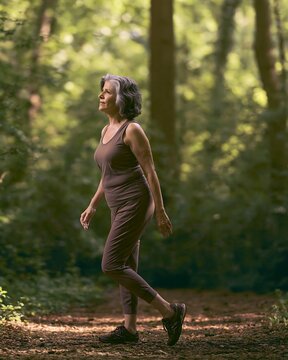 Mature woman walking on a sunlit forest path, active menopause lifestyle concept for women&rsquo;s health, grounding, mental wellbeing, healthy aging and outdoor wellness content