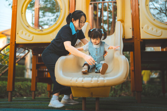 Mother teaching toddler daughter to use playground slide outdoors, gently supporting child during active play, early learning, parental guidance, safety, confidence,
