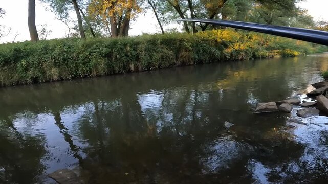 POV of an angler holding a long fishing pole rod over a calm forest river. First person view of summer freshwater angling.