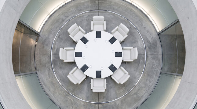 Top view of a modern circular conference room with a white round table and eight white chairs arranged symmetrically on a grey concrete floor