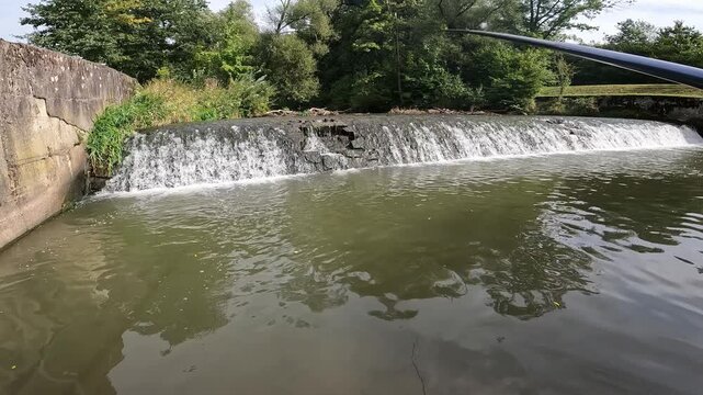 Fishing rod tip over a river weir with cascading water. Angling near a small dam waterfall in a summer green forest environment.