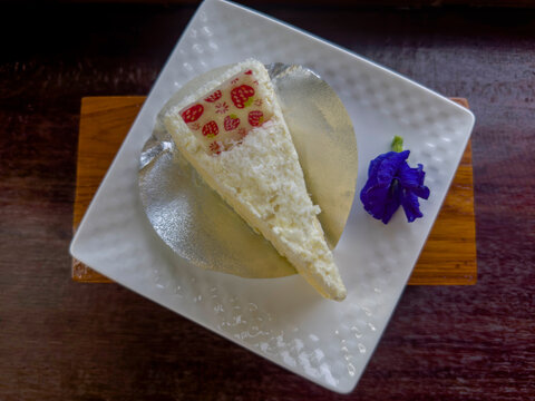 top view sweet triangular slice of white cake resting on a circular metallic silver paper tray and butterfly pea flower on a white ceramic plate on wooden background, food, dessert, copy space