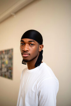 A focused portrait of a young African American man wearing a black durag and a white t-shirt, standing indoors against a neutral wall while looking directly at the camera with calm confidence.