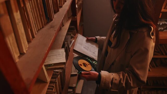 Woman examines an old archival record among crowded wooden shelves in warm dim light. Concept historical research, preservation, publishing and documentary content.