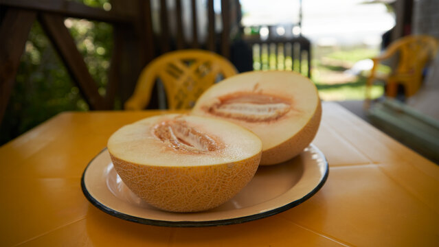 A ripe melon cut in half, sitting on a large plate on a table on the terrace of a summer house. Droplets of juice are visible on the cut surface.