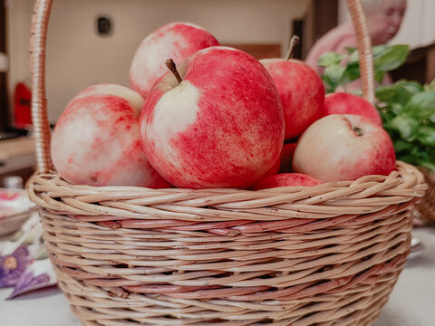 Basket of red and white apples with rustic wicker texture, leafy branch, and warm indoor background, arranged as a harvest still life on a table