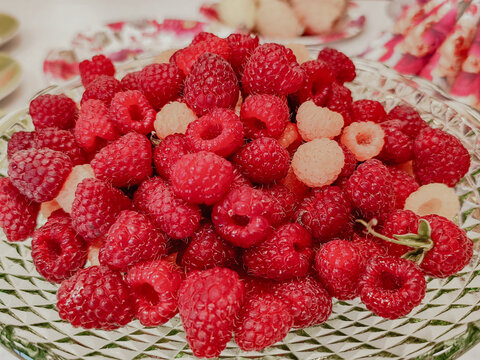 Fresh red and pale raspberries piled in a clear textured glass dish with floral tableware and pink accents, close-up food still life on a dining table