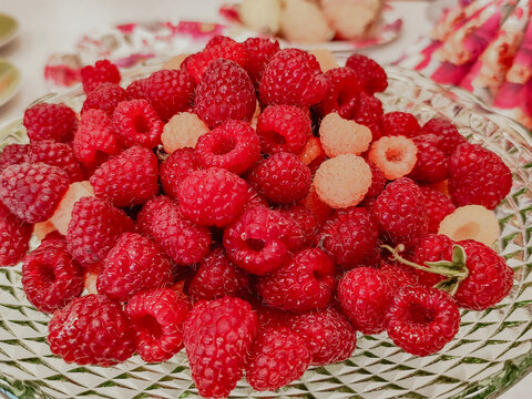 Fresh red and pale raspberries piled in a clear patterned glass serving bowl with scattered berries and floral tableware in a bright still life