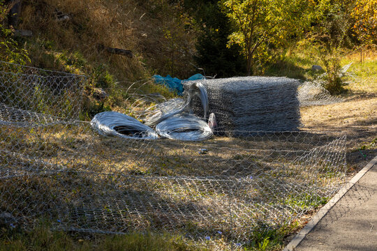 Rolls of galvanized hexagonal wire mesh and gabion mesh stored outdoors near a wooded area, showing construction and landscaping materials in a natural environment.