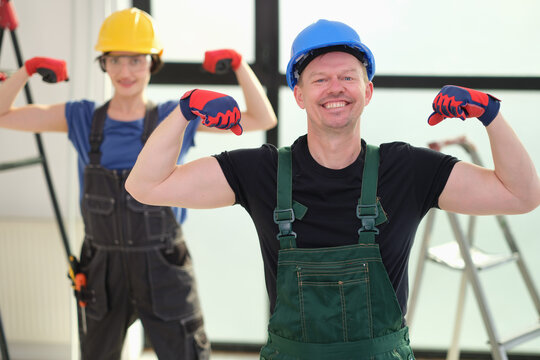 Cheerful workers wearing safety helmets and gloves pose with flexed arms in sunlit interior, with ladders and tools in background, suggesting renovation, teamwork, strength, and skilled labor