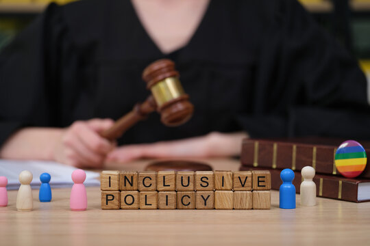 A judge in a black robe holds a gavel, presiding over a discussion on inclusive policy. Colorful wooden figures surround the words 'INCLUSIVE POLICY' on the table, representing diverse viewpoints.