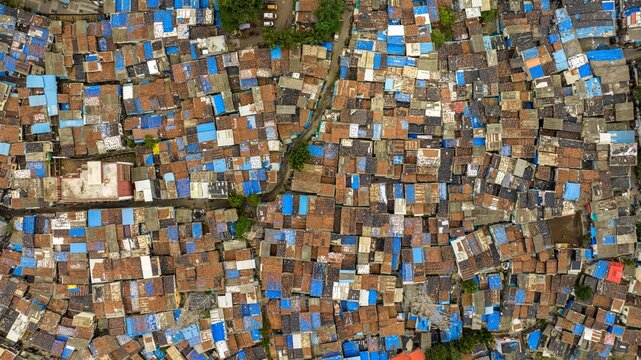 Aerial view of the dense urban fabric of Dharavi Slums, featuring a complex network of rooftops with blue tarpaulins and brown tiles, and narrow alleyways in Mumbai, Maharashtra, India.