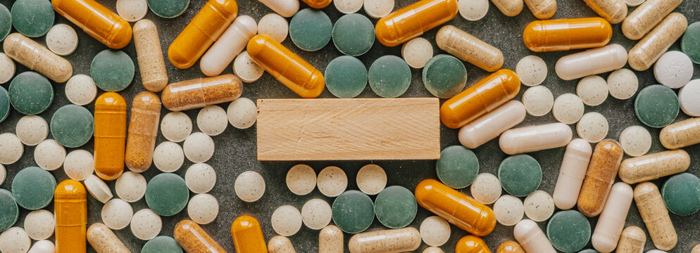 Top down view of wooden block copy space framed by various vitamin pills and colorful supplement capsules on dark surface
