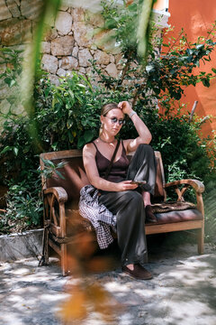 Young woman using smartphone in cozy Mediterranean cafe and garden courtyard, showing solo travel, vacation lifestyle, leisure, mobile connection and relaxed summer tourism.
