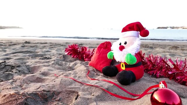Santa Claus plush toy sits with red Christmas decorations on a sandy beach, celebrating the holidays in a warm tropical location with the ocean in the background during sunrise