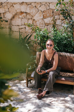 Young woman using smartphone in cozy Mediterranean cafe and garden courtyard, showing solo travel, vacation lifestyle, leisure, mobile connection and relaxed summer tourism.