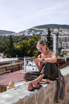Young woman using smartphone in cozy Mediterranean cafe and garden courtyard, showing solo travel, vacation lifestyle, leisure, mobile connection and relaxed summer tourism.