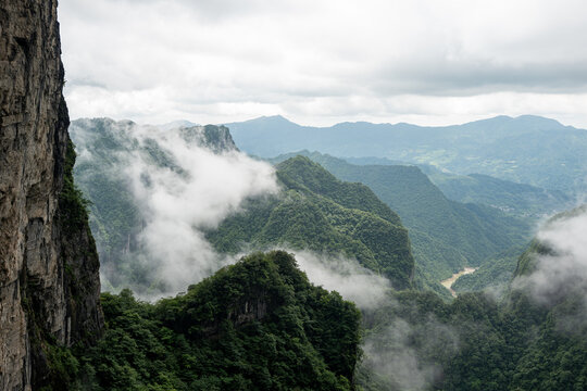 Sweeping natural panorama showcasing towering rocky precipices alongside dense, lush woodlands. Soft white clouds roll through the deep ravine, revealing a distant muddy river under a cloudy sky.