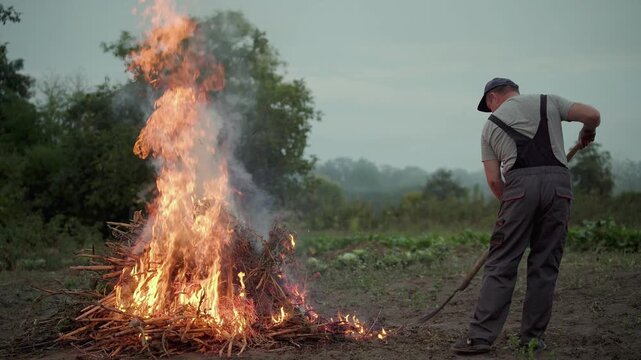 Farmer uses a long handled tool or fork to tend burning branches as flames rise and smoke drifts across a rural field, with trees and hazy hills visible in the background