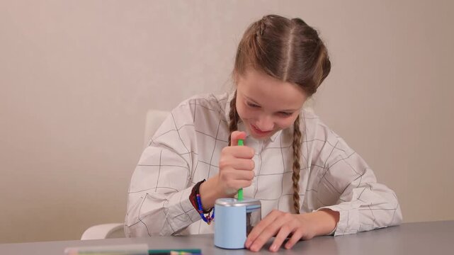Hands rotating crank to sharpen. Focused student sharpening pencil with colorful accessories visible. Student attentively sharpening green pencil using hand crank on desk