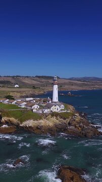 Aerial perspective of Pigeon Point Lighthouse standing tall on a rugged rocky headland along the Pacific Ocean coastline in California, surrounded by rolling coastal hills and ocean swells.