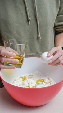 Person in green hoodie prepares ingredients for baking, holding an egg and glass of oil over a red mixing bowl filled with flour and egg yolks