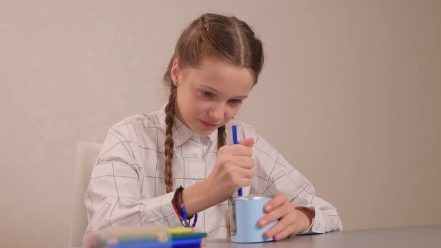 Hands rotating crank to sharpen. Focused student sharpening pencil with colorful accessories visible. Student attentively sharpening green pencil using hand crank on desk