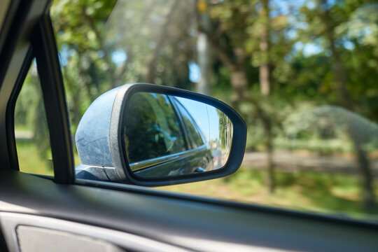 Car side mirror reflects vehicle body and road behind with green trees outside