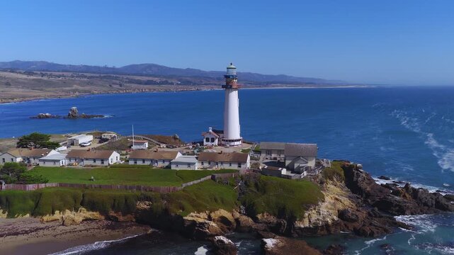 Aerial view moves toward Pigeon Point Lighthouse perched on rocky cliffs along the California coastline, surrounded by breaking ocean waves and coastal terrain.