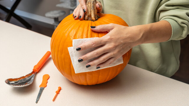Person cleaning a bright orange pumpkin with a paper towel on a light-colored table, carving tools arranged nearby for Halloween decoration preparation