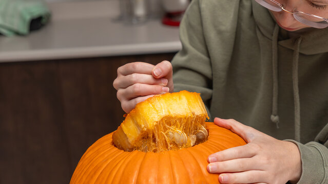 Child carefully removes the top of a bright orange pumpkin for Halloween decoration in a kitchen setting with countertop and utensils visible in the background