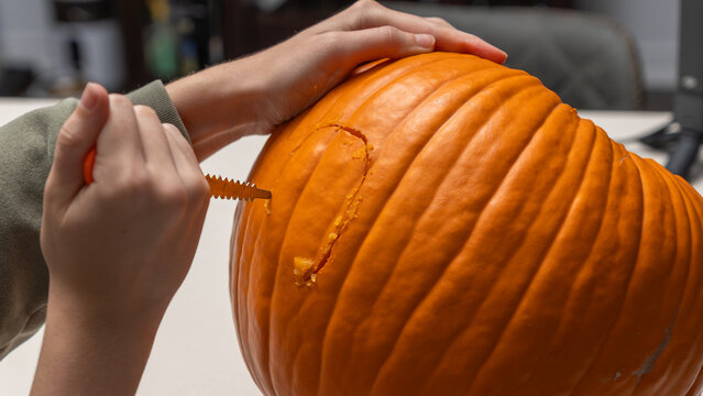 Hands carving a pumpkin for Halloween with a serrated knife on a white table, workspace visible in the background