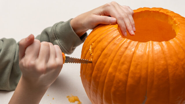 Hands of a person carving an orange pumpkin for Halloween with a small saw tool on a light-colored surface, showcasing the intricate details of the pumpkin's texture