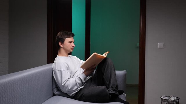 Young man relaxed on a modern sofa, absorbed in a book, enjoying a quiet evening of peaceful solitude and calm reading downtime at home