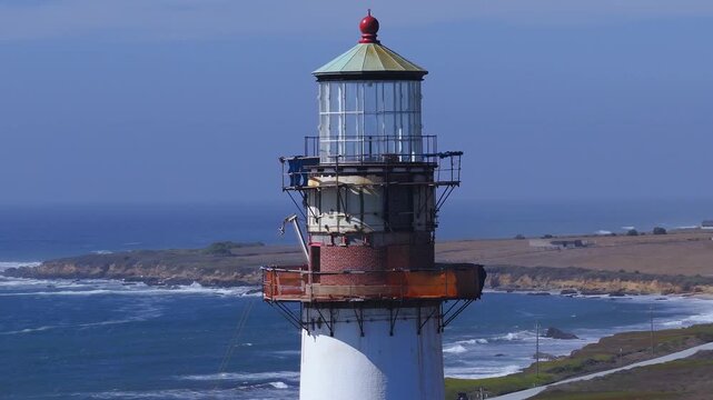 Aerial view shows the upper tower and lantern room of Pigeon Point Lighthouse under restoration along the Pacific coast of California, with waves breaking on the rocky shoreline in daylight.