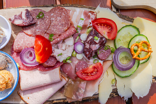 Traditional Austrian Bavarian Brettljause alpine snack board served on a wooden platter, with cured meats, sausages, cheeses, tomato, cucumber, red onion and mini pretzel