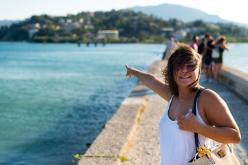 A smiling woman stands on a pier and points toward the sea in Kerkira, Greece. Green hills and coastal buildings are visible across the water in the background. © dsbedarieva