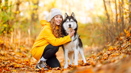 Young woman in yellow raincoat walks with husky through an autumn forest. Woman enjoys moment with pet, surrounded by yellow leaves at sunset. A concept of friendship and walking. © maxbelchenko