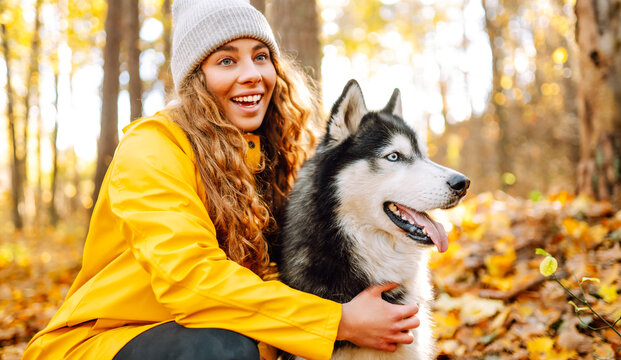 Young woman in yellow raincoat walks with husky through an autumn forest. Woman enjoys moment with pet, surrounded by yellow leaves at sunset. A concept of friendship and walking.