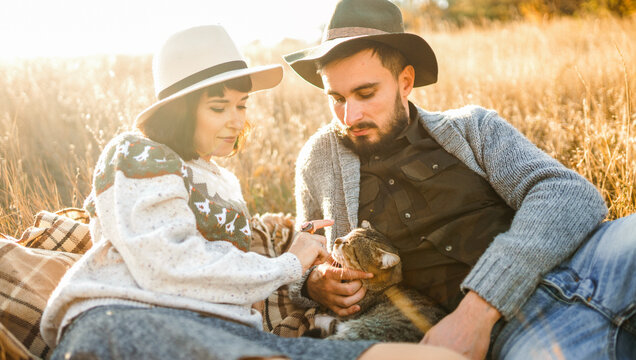Young couple spends time together sitting on blanket in field, petting a cat. Couple enjoys relaxing outdoors. Concept of love, enjoyment, and day off.