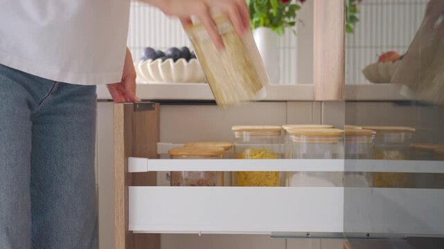 Woman opens kitchen pantry drawer, takes jar with oats and closes storage unit. Concept of food organization, pantry access and everyday kitchen routine