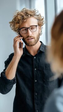 Young man with curly hair and glasses talking on a smartphone indoors. Casual professional wearing a dark shirt engaged in a phone conversation in a modern setting