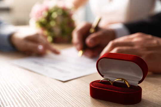 Notary helping couple with signing of marriage contract and wedding rings in box on wooden table indoors, selective focus