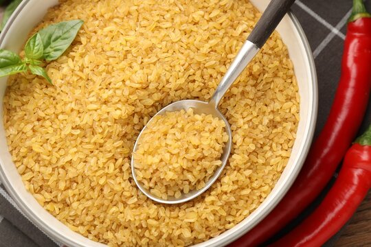 Raw bulgur in bowl, spoon, basil and chili peppers on wooden table, flat lay