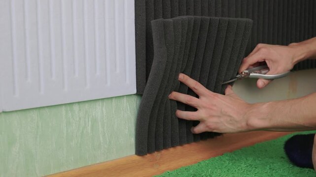 Cutting and shaping acoustic foam for soundproofing purposes. Meticulously trimming black foam with utility knife to ensure snug fit around baseboards and flooring edges