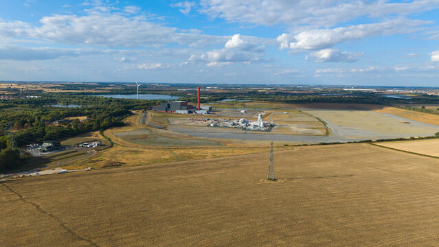 Aerial view of Millbrook Power Plant with its red chimney, surrounded by open fields, industrial infrastructure, and distant lakes in Stewartby, England, United Kingdom.