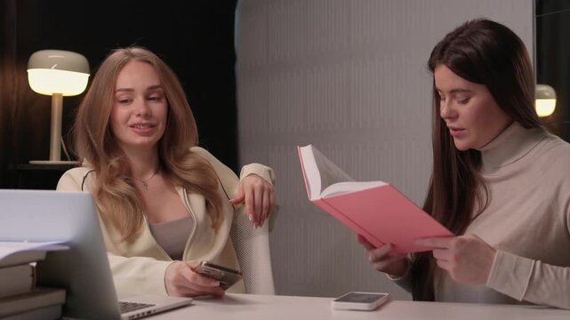 Two females engaged in studying. Women collaborating at desk with books and laptop in office. Two women focused on reading and notetaking in contemporary work environment