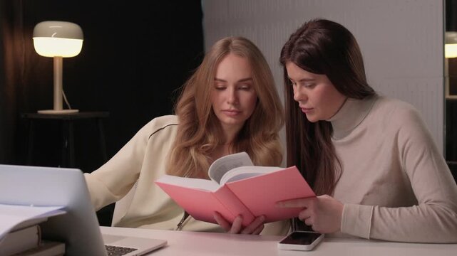 Two females engaged in studying. Women collaborating at desk with books and laptop in office. Two women focused on reading and notetaking in contemporary work environment