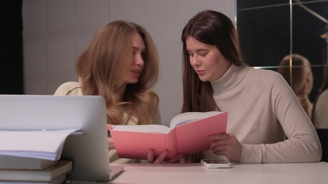 Two females engaged in studying. Women collaborating at desk with books and laptop in office. Two women focused on reading and notetaking in contemporary work environment
